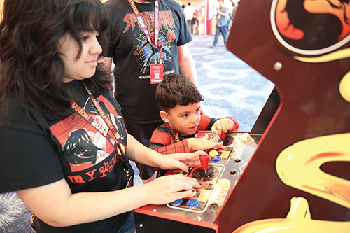 Woman and a young child play a colorful arcade game with buttons and joystick at a gaming area.