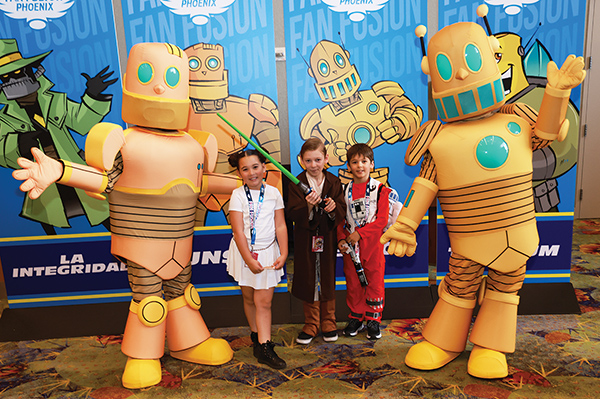 Three kids posing with four large orange robot mascot characters at a convention photo booth.