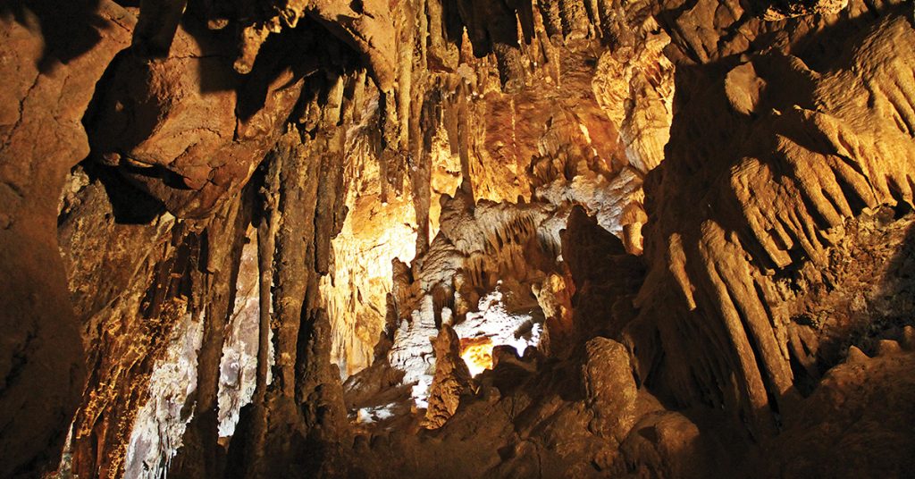 Inside a large cave with tall stalactites hanging from the ceiling and stalagmites rising from the floor, lit by warm golden lights.