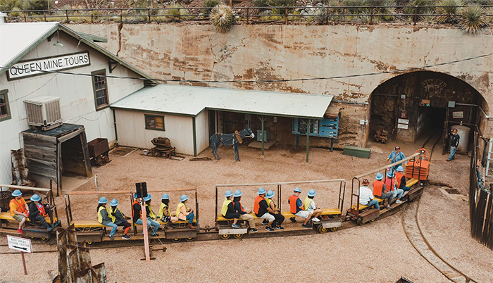 Visitors wearing blue hard hats ride a small mine train into a tunnel at the Queen Mine Tours entrance.