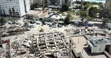 Aerial view of a city block with a completely collapsed building reduced to rubble, surrounded by rescue vehicles and workers.