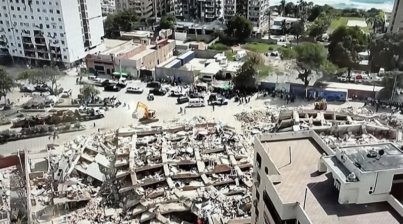 Aerial view of a city block with a completely collapsed building reduced to rubble, surrounded by rescue vehicles and workers.