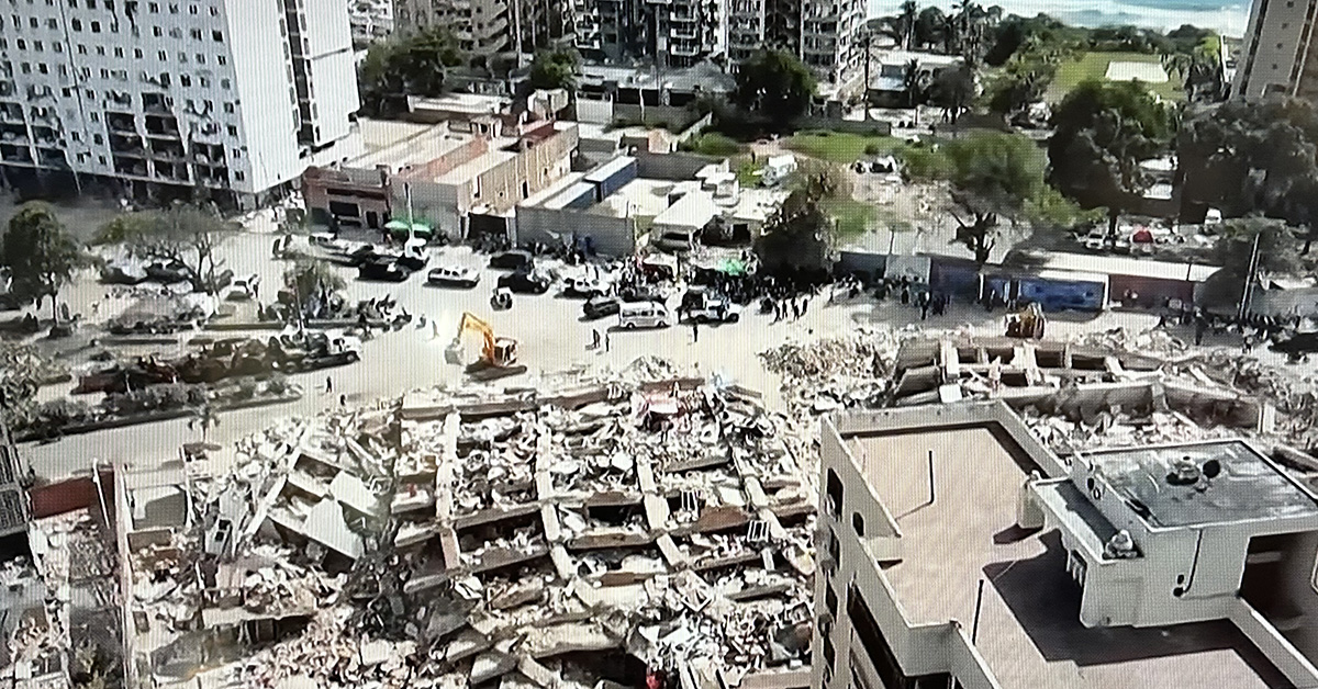 Aerial view of a city block with a completely collapsed building reduced to rubble, surrounded by rescue vehicles and workers.