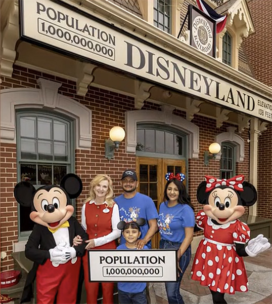 A family poses with Mickey and Minnie Mouse at Disneyland holding a sign that reads 'Population 1,000,000,000.