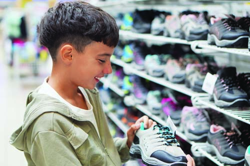 Smiling boy holding a sneaker while shopping in a shoe store with rows of shoes on shelves behind him.