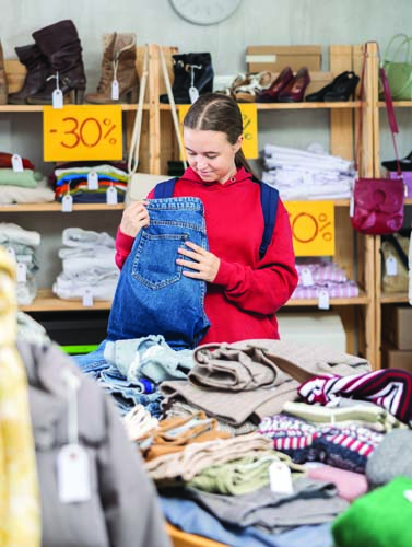 Teenage girl in a red hoodie holding up a pair of jeans while shopping at a store with sale signs.