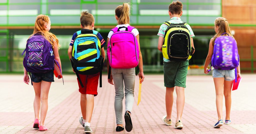 Five kids walking toward a school building from behind, each wearing a colorful backpack.
