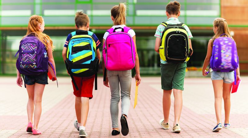 Five kids walking toward a school building from behind, each wearing a colorful backpack.