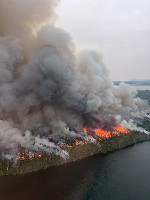 Aerial view of a massive wildfire burning a forest next to a lake, with huge orange flames and thick gray smoke filling the sky.
