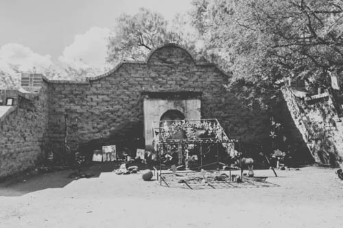 Black-and-white photo of the El Tiradito shrine: an old stone wall with an arched doorway. People have left flowers, pictures, and gifts in front of it.
