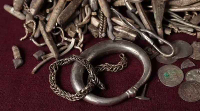 A pile of old silver treasure on red cloth: a thick ring, a braided chain, twisted wire, small bars, and worn coins with faded writing.